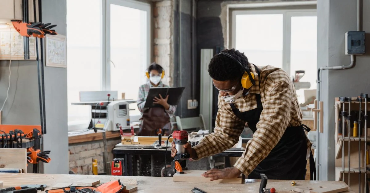 Charpentier utilisant des outils dans un atelier avec un collègue en arrière-plan.